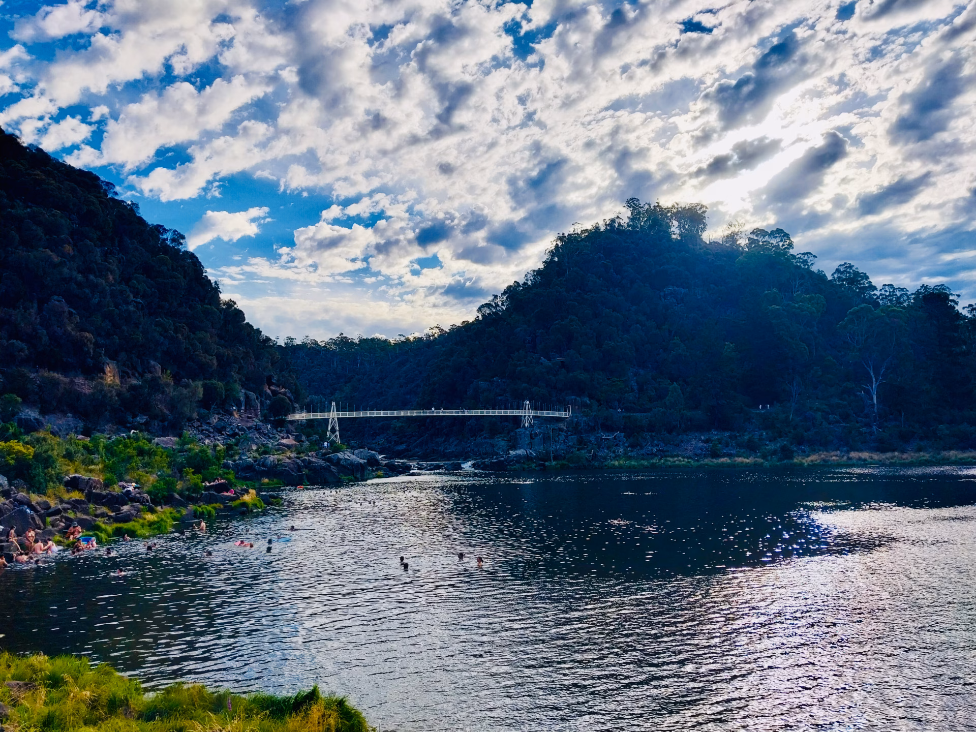 Natural gorge at Cataract Gorge Launceston with people swimming in the cold dark water and Alexandra Suspension Bridge visible in the background under a dramatic cloudy sky
