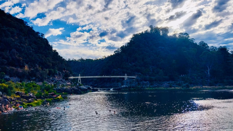 Natural gorge at Cataract Gorge Launceston with people swimming in the cold dark water and Alexandra Suspension Bridge visible in the background under a dramatic cloudy sky