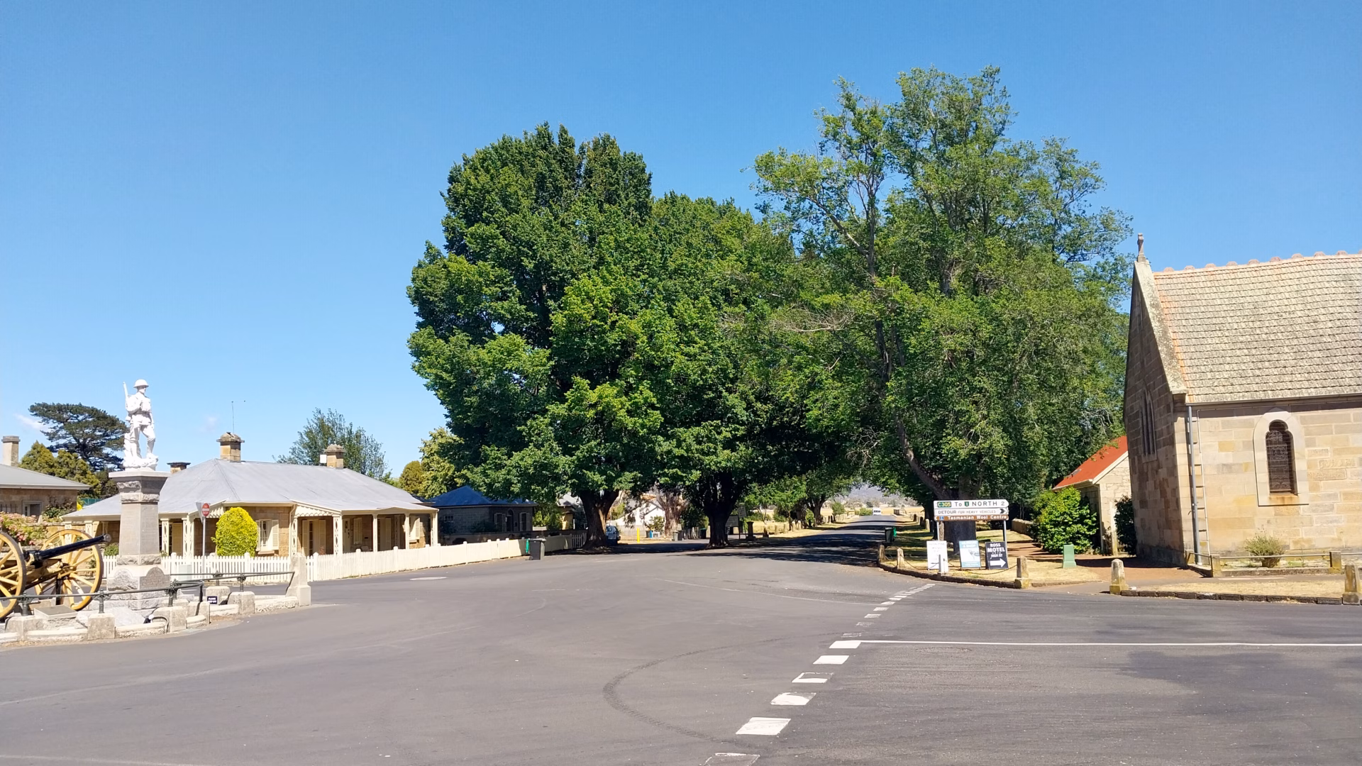 Looking down Church Street in Ross Village Tasmania with the war memorial and cannon on the left the sandstone church on the right and large elm trees lining the empty road under a perfect blue sky