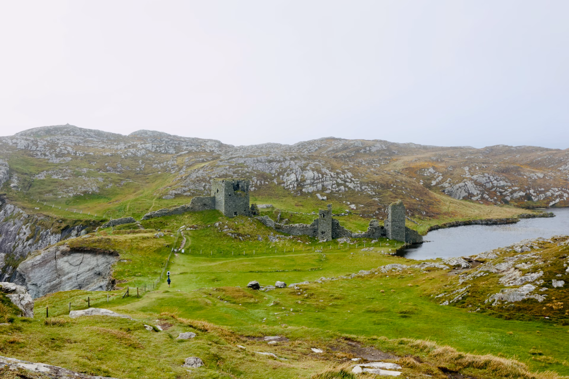Full Three Castle Head castle complex from elevation — three towers, connecting walls, tiny lone hiker, Lough Fadda to the right