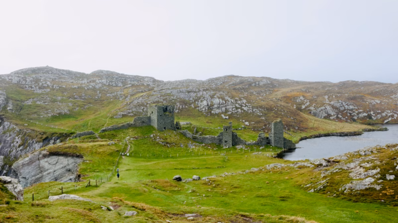 Full Three Castle Head castle complex from elevation — three towers, connecting walls, tiny lone hiker, Lough Fadda to the right
