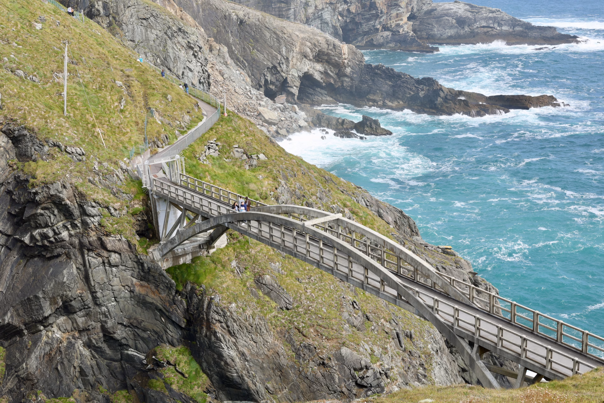 Mizen Head Bridge spanning the gorge from above with people crossing and turquoise Atlantic below