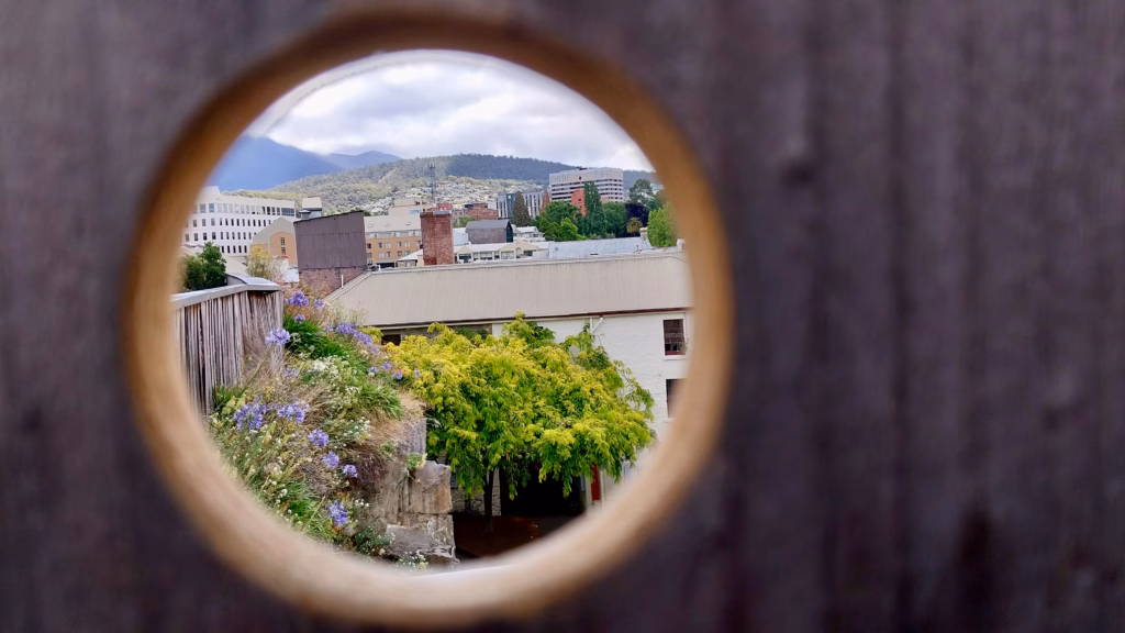 View of Hobart rooftops and Mount Wellington through a circular hole in a wooden fence at the top of Kelly's Steps