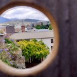 View of Hobart rooftops and Mount Wellington through a circular hole in a wooden fence at the top of Kelly's Steps