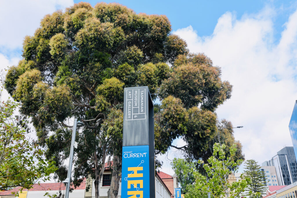 TMAG signpost under a eucalypt tree with a Hobart Current exhibition banner