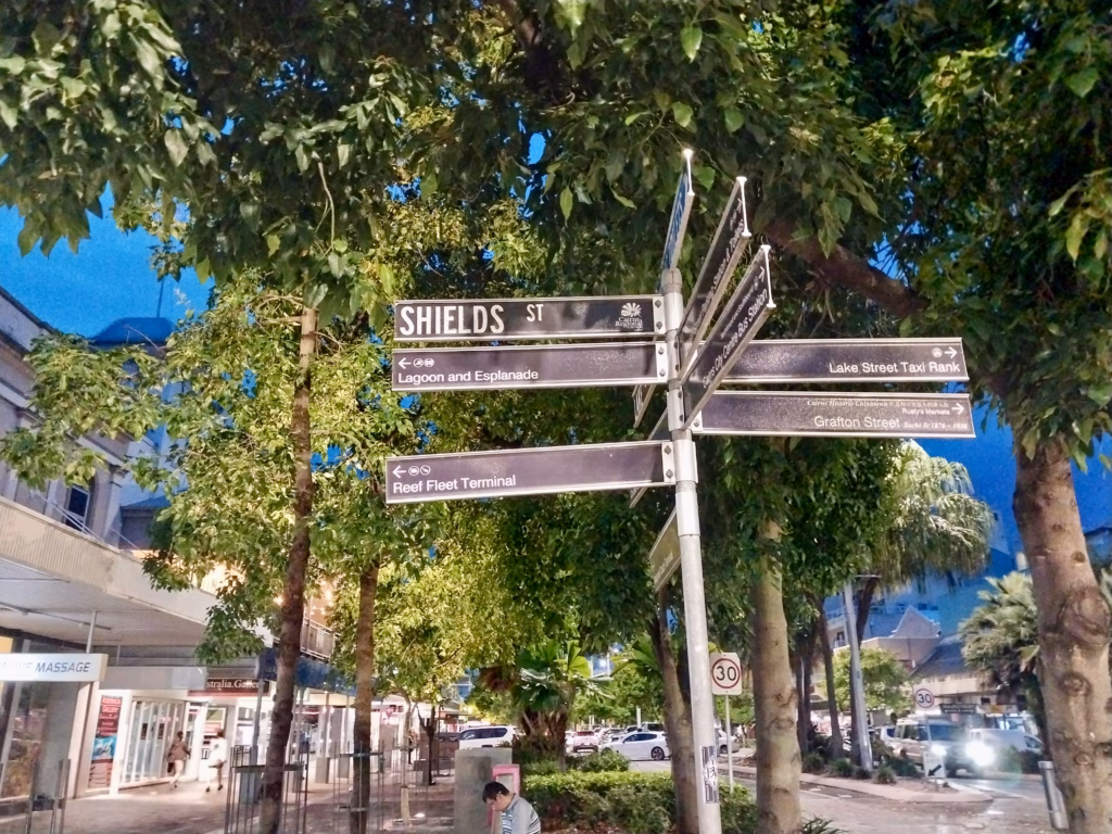 A directional signpost on Shields Street at night, pointing to the Lagoon, Esplanade, Reef Fleet Terminal, Lake Street Taxi Rank, and Cairns' historic Chinatown