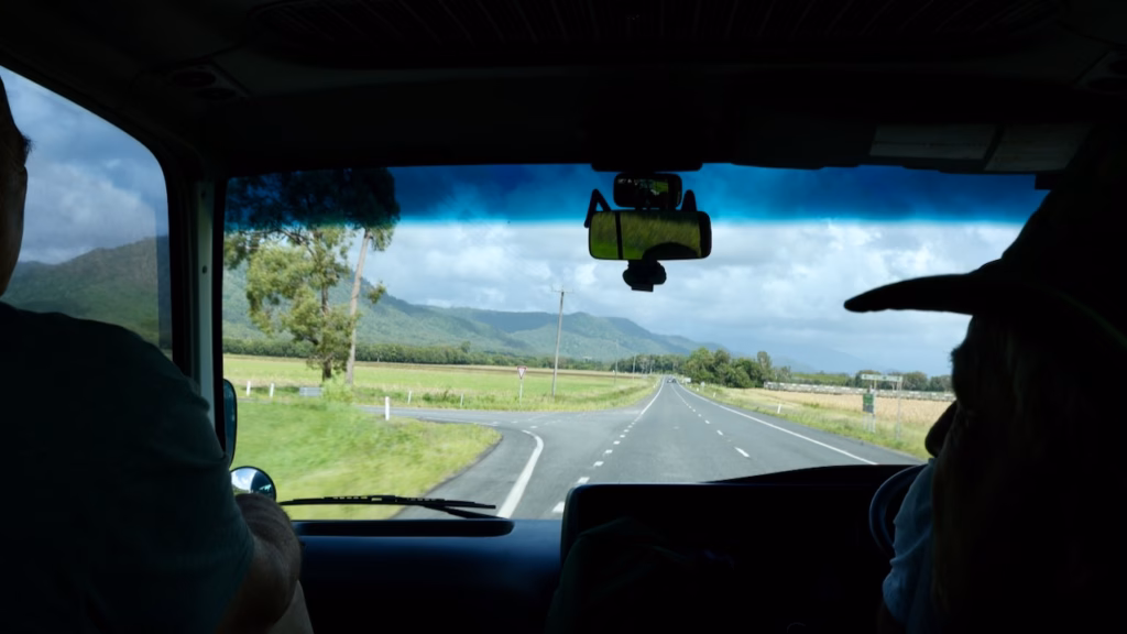View through the tour vehicle's windscreen along an open highway lined with sugarcane fields and distant mountains