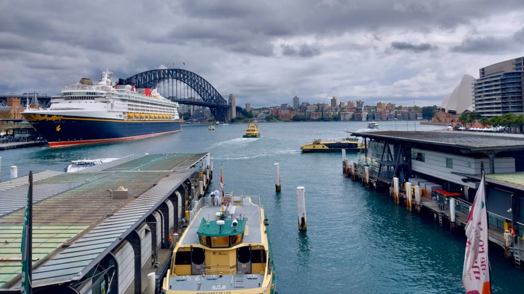 Elevated view down over Circular Quay's ferry wharves, the Margaret Olley ferry docked in the foreground, a cruise ship and the Harbour Bridge behind, Opera House visible far right
