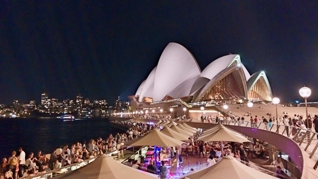 Elevated night view over a packed outdoor dining and bar precinct at Circular Quay, Sydney Opera House lit up behind the tables