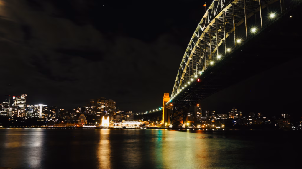 Sydney Harbour Bridge arching toward Luna Park's lit ferris wheel across the water at night, city skyline on the left, from the Dawes Point area