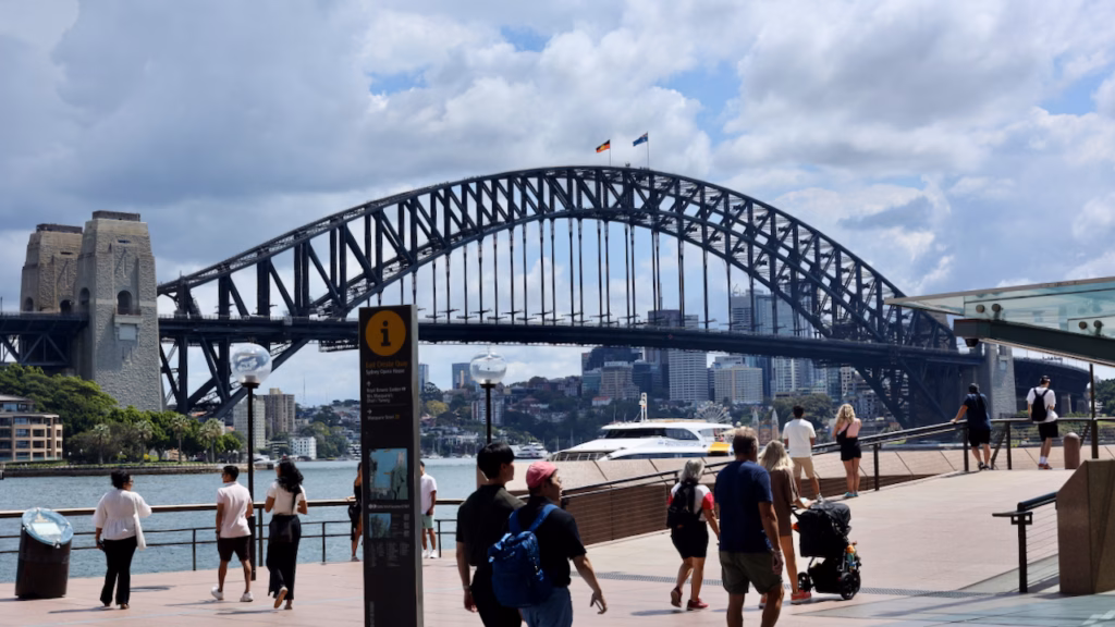 Sydney Harbour Bridge behind a busy pedestrian promenade at East Circular Quay, wayfinding signpost for Royal Botanic Garden and Mrs Macquarie's Chair, a ferry passing under the bridge