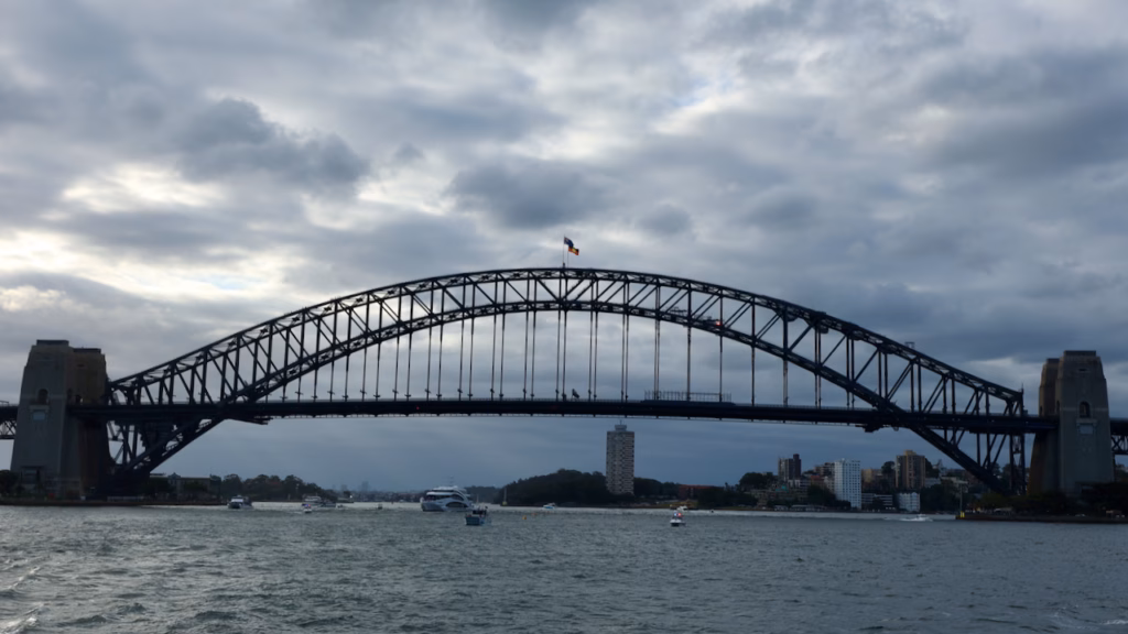 Sydney Harbour Bridge from the water on an overcast day, boat railing visible in the foreground, both pylons and the full arch in frame