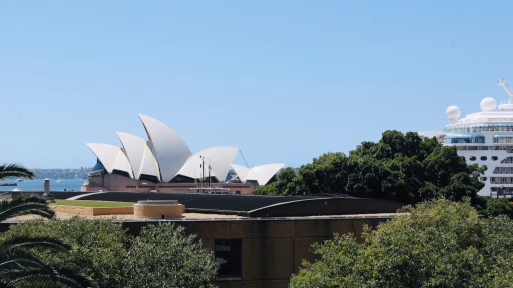 Sydney Opera House sails glimpsed through trees from near Observatory Hill, a cruise ship at the Overseas Passenger Terminal visible alongside