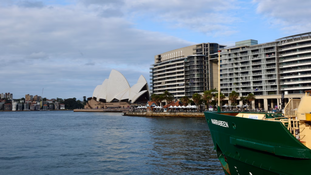 Sydney Opera House and apartment buildings viewed from the water, the bow of a Sydney ferry named Narrabeen visible in frame