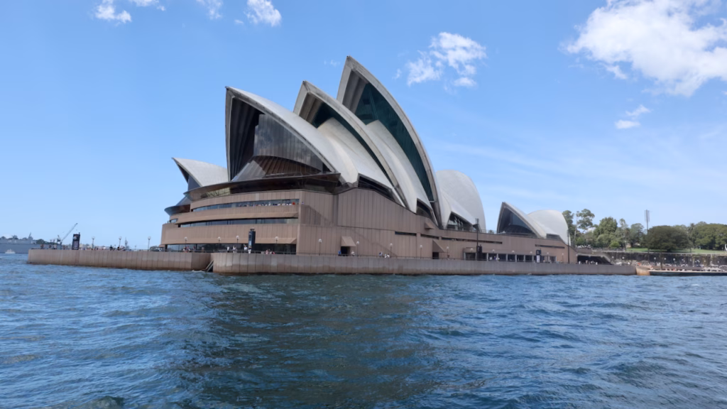 Sydney Opera House seen from the ferry rounding Bennelong Point, with a grey navy ship at Garden Island on the left