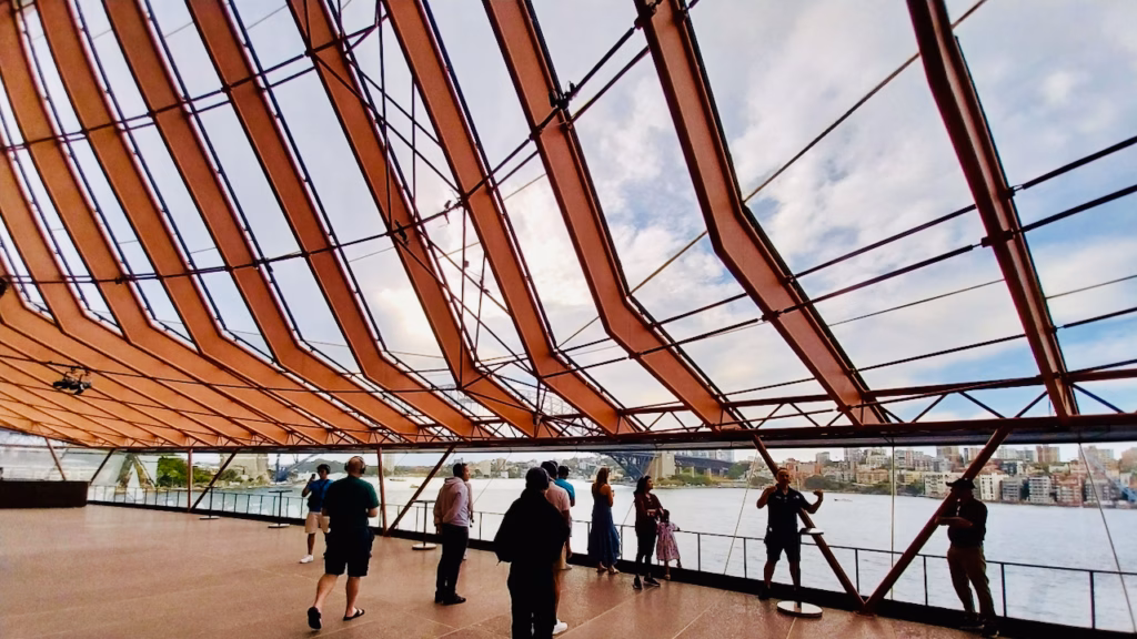 A guided tour group standing inside the Sydney Opera House's glass-walled colonnade, the tour guide gesturing while explaining, Sydney Harbour and the Harbour Bridge visible through the glass