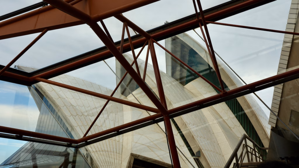 Sydney Opera House sail shells viewed through an orange-painted steel roof truss from inside a glass-enclosed walkway during the guided tour