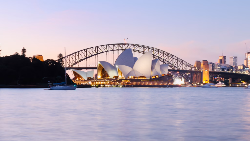 Sydney Opera House and Harbour Bridge in stacked framing from inside the Royal Botanic Garden at dusk, pink sky, a treed point of land and moored sailboat in the foreground