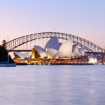 Sydney Opera House and Harbour Bridge in stacked framing from inside the Royal Botanic Garden at dusk, pink sky, a treed point of land and moored sailboat in the foreground
