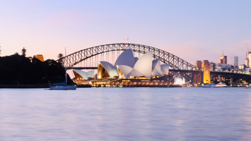 Sydney Opera House and Harbour Bridge in stacked framing from inside the Royal Botanic Garden at dusk, pink sky, a treed point of land and moored sailboat in the foreground