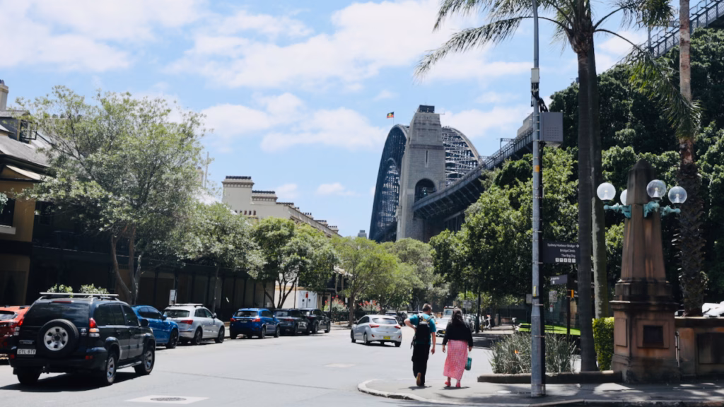 Street-level view in The Rocks with palm trees, a Sydney Harbour Bridge and BridgeClimb signpost, and the bridge visible in the background