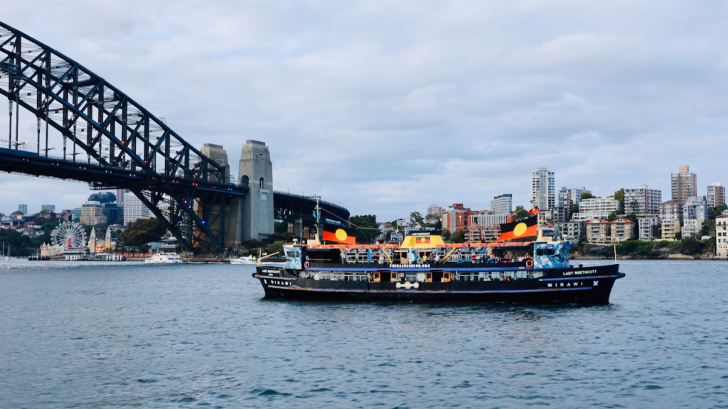 The Wirawi, a black-hulled Aboriginal-owned harbour cruise vessel formerly the Lady Northcott ferry, passing the Sydney Harbour Bridge's northern pylon with Aboriginal flags flying