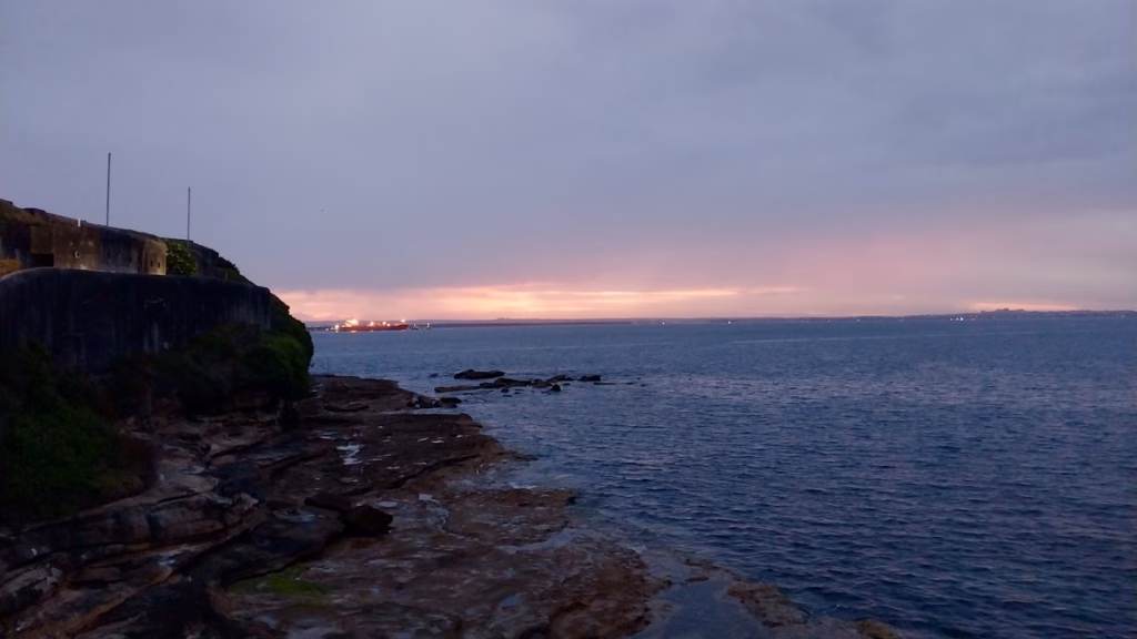 Pink afterglow on the horizon over Botany Bay, with the walls of Bare Island Fort and dark rock shelves on the left