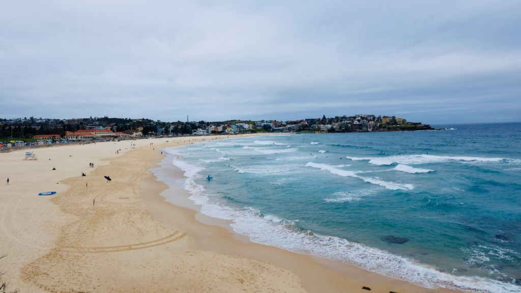 Bondi Beach seen from the path above the south end, with surfers in the water and the North Bondi headland across the bay