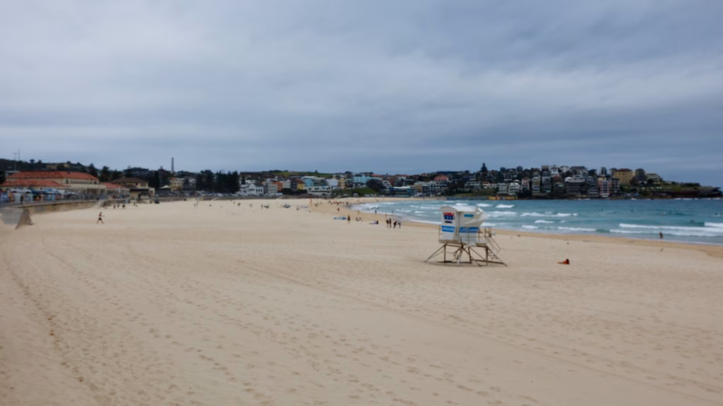A lifeguard tower on a nearly empty Bondi Beach under heavy grey cloud, with the Bondi Pavilion on the left