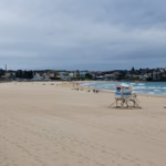 A lifeguard tower on a nearly empty Bondi Beach under heavy grey cloud, with the Bondi Pavilion on the left