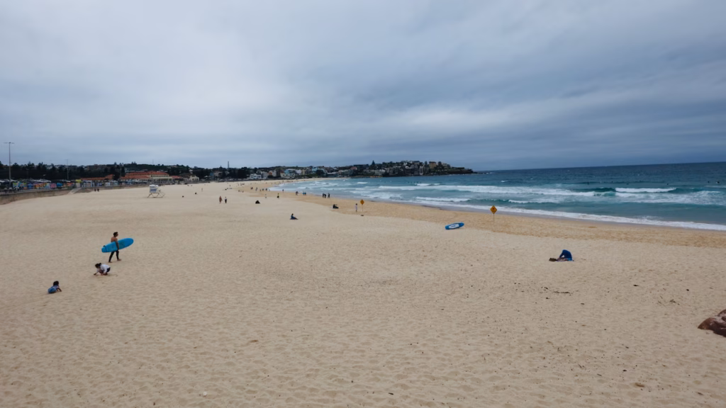 A surfer carrying a blue surfboard across Bondi Beach while children dig in the sand