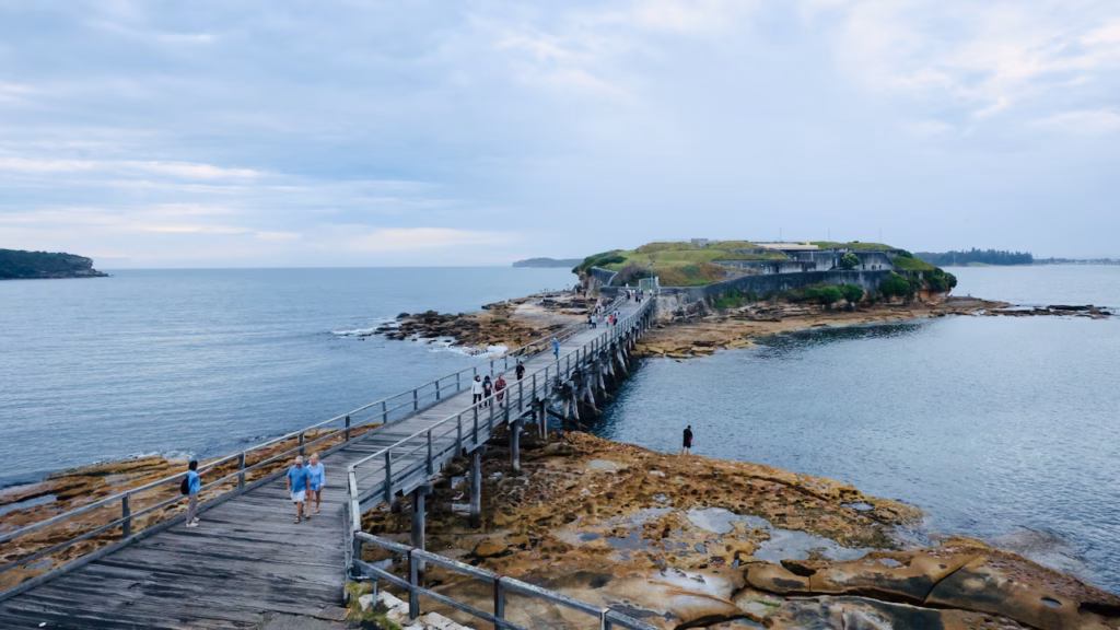 People sitting on folding chairs on the grassy La Perouse headland, looking out over Bare Island and Botany Bay under a cloudy sky