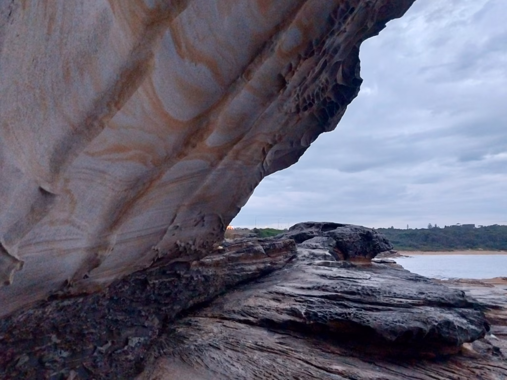 Close view of a curved sandstone overhang with orange stripes and honeycomb weathering, framing the bay behind