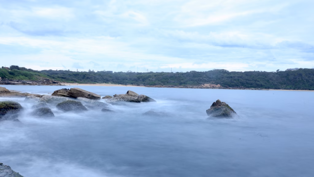 Long-exposure photo of water flowing smoothly around rocks off La Perouse, with the wooded far shore of Botany Bay behind