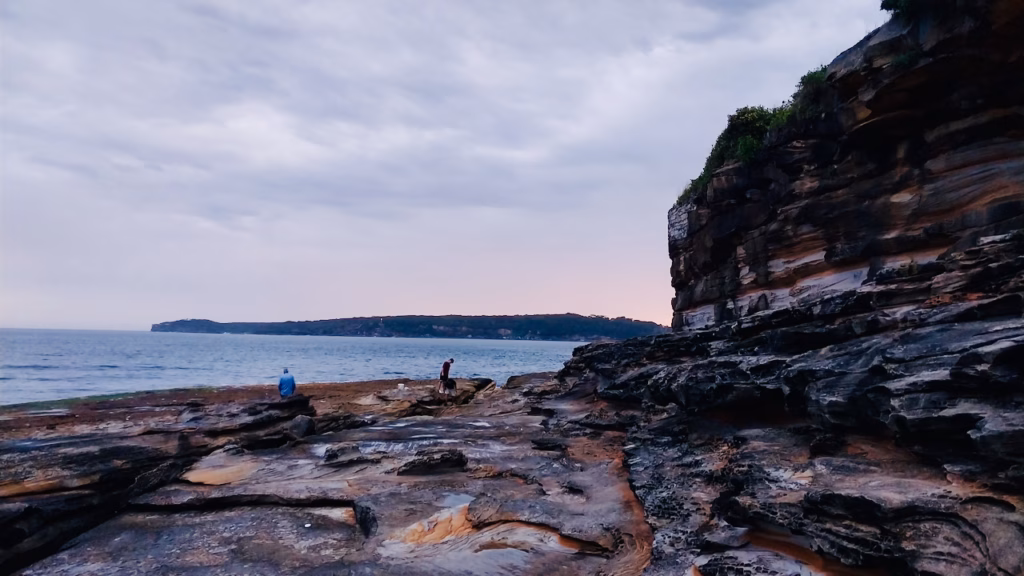 Two people fishing from a dark sandstone rock shelf below layered cliffs at La Perouse, with a headland across the water