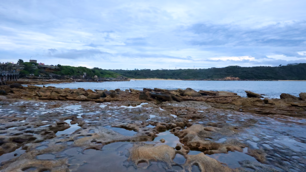 Flat sandstone rock platform full of shallow rock pools at La Perouse, with the headland and a beach in the distance