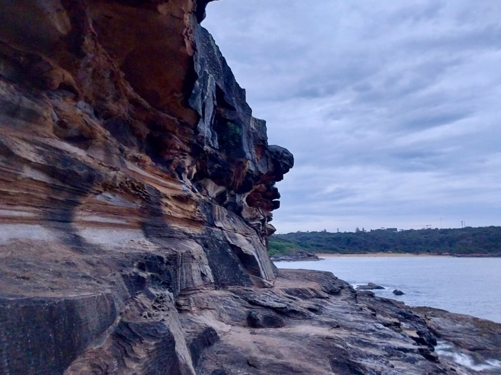 Layered sandstone cliff overhang with orange and grey bands above a rock shelf, with Botany Bay and a beach beyond