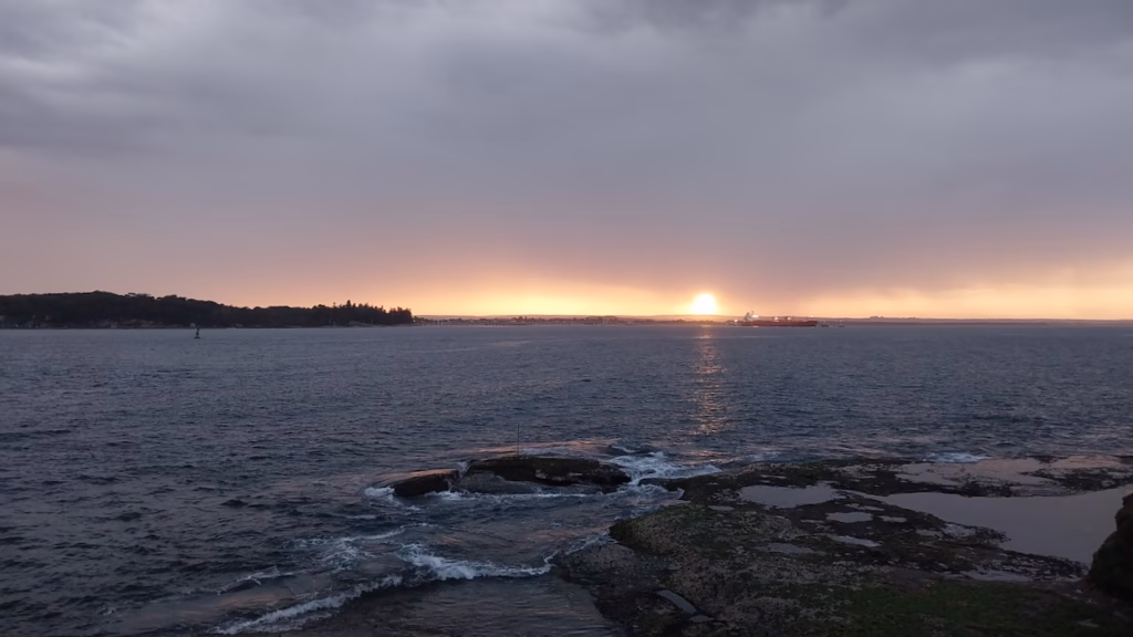 Sun setting through a gap in heavy cloud over Botany Bay, with its reflection on the water and rock shelves in the foreground at La Perouse