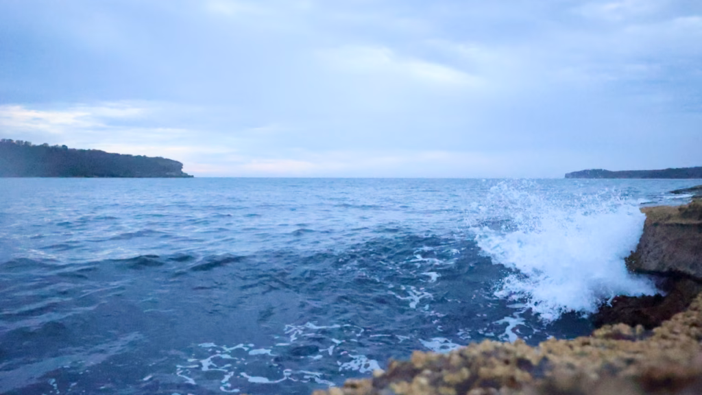 Wave breaking against the edge of a rock shelf at La Perouse, sending white spray into the air, with headlands across the bay